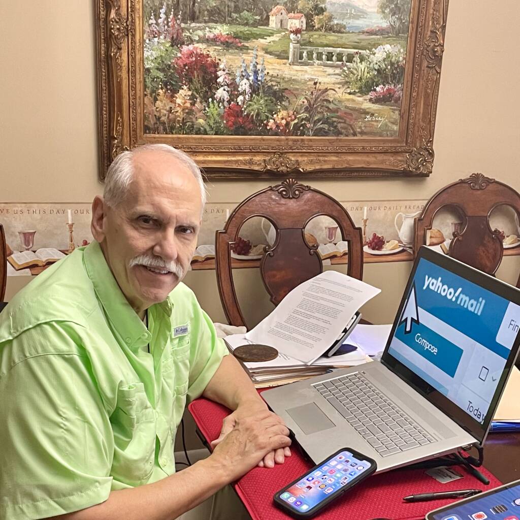 A man sits at a table and faces the camera. There is a laptop, a smartphone, tablet computer and various papers on the table.