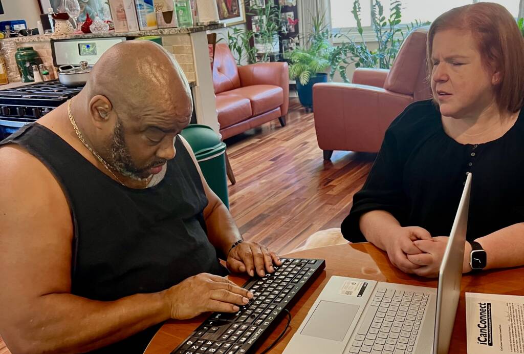 Larry (left) and iCanConnect trainer Lisa (right) sit at a table during a training session. Larry is using his laptop and braille display, while Lisa looks on, ready to assist.