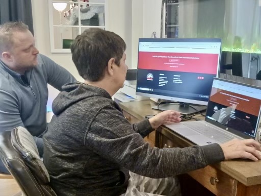 A woman sits at a desk and uses a laptop computer and big screen monitor on the desk in front of her. A man sits next to her and looks at the large screen monitor.