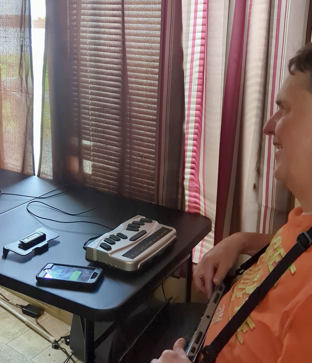 A man sits at a desk and uses a braille display and smartphone.