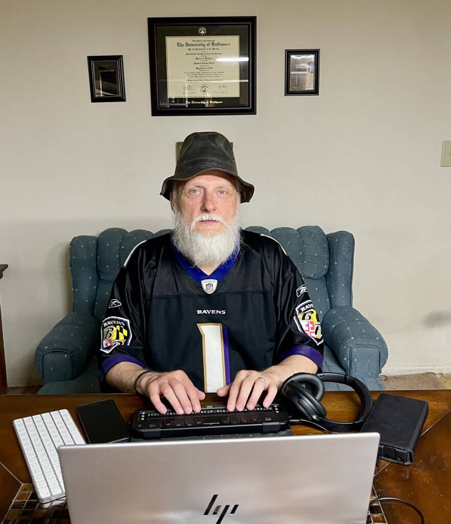 An older man with a white beard and a dark bucket hat is seated at a desk with a laptop, keyboard, braille display, and headphones. He is wearing a black Baltimore Ravens football jersey, and behind him on the wall are three framed documents.