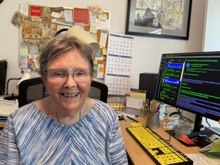 An elderly woman with short, gray hair and glasses smiles while sitting at a computer desk with a large monitor displaying green text on a dark background.