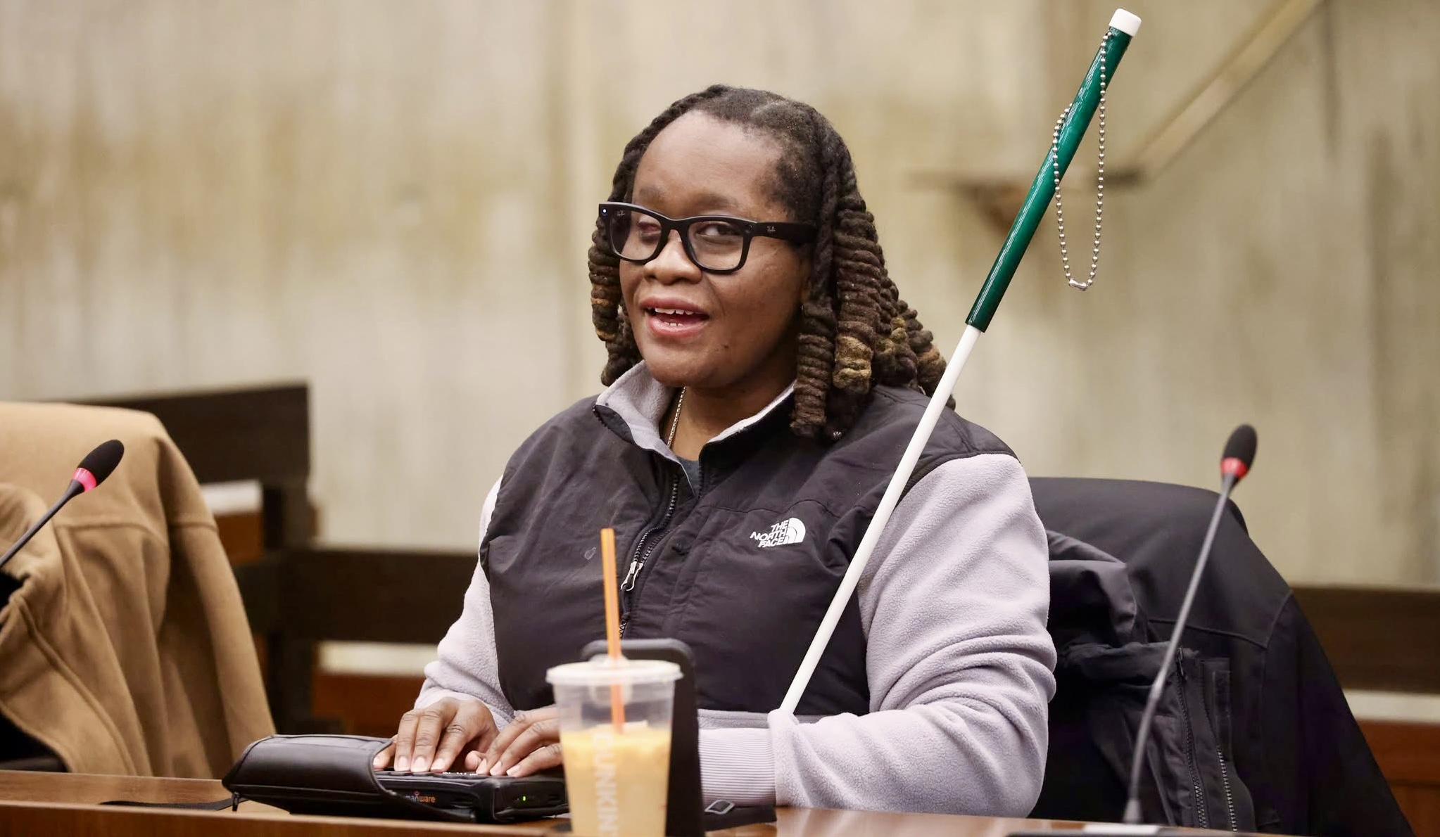 A image of Casandra Xavier, a Black woman with long locs and black-rimmed glasses, sitting at a long wooden table in a professional setting. She is wearing a grey and black North Face fleece vest. Her hands are resting on a refreshable braille display, and a white cane with a green handle leans against her shoulder. Microphones are visible on the table, suggesting a public speaking or board meeting environment.