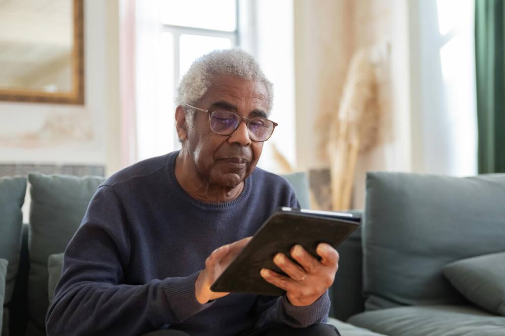 An older man with gray hair and glasses sitting on a sofa while using a tablet.