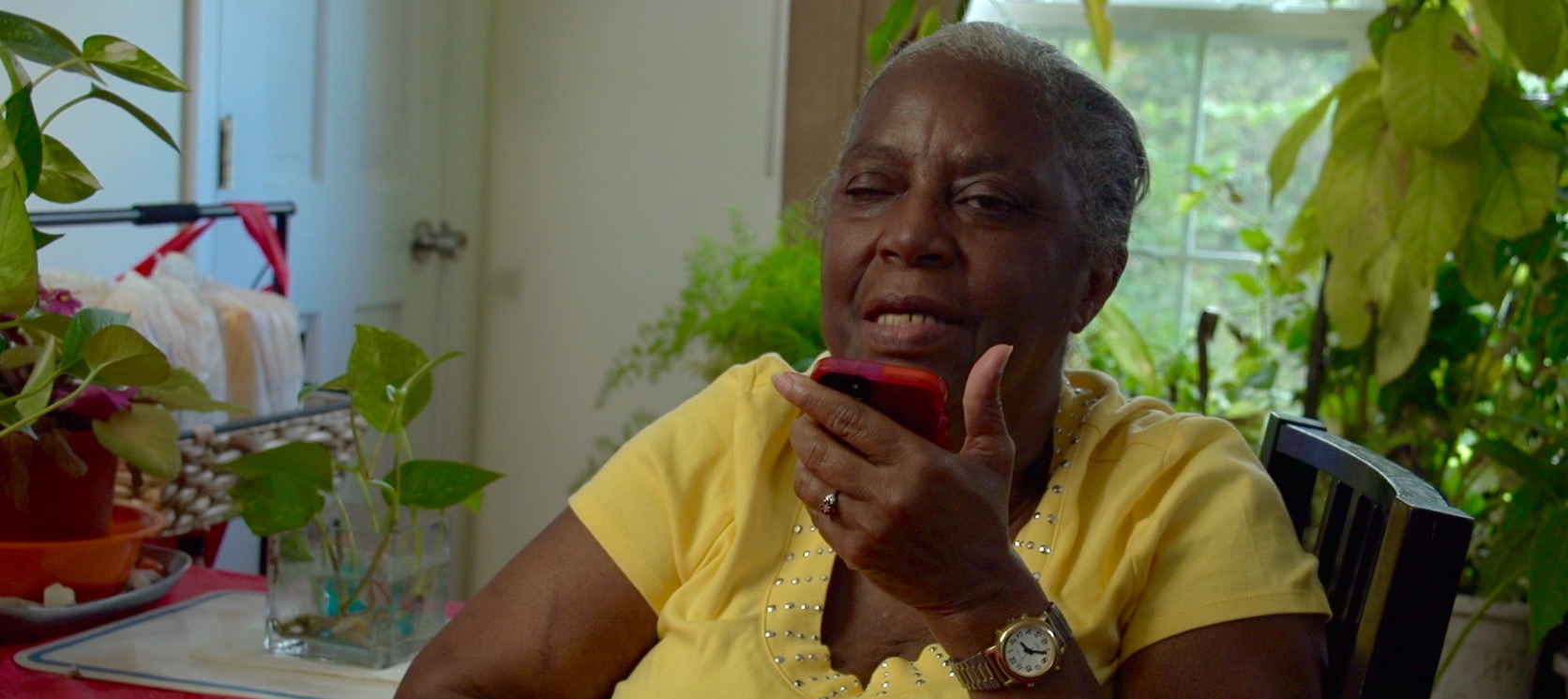 Annette Rogers, an older Black woman wearing a bright yellow shirt and a watch, sits indoors surrounded by lush green houseplants. She is holding a red smartphone close to her face, appearing to speak into the microphone or listen intently to the speaker.