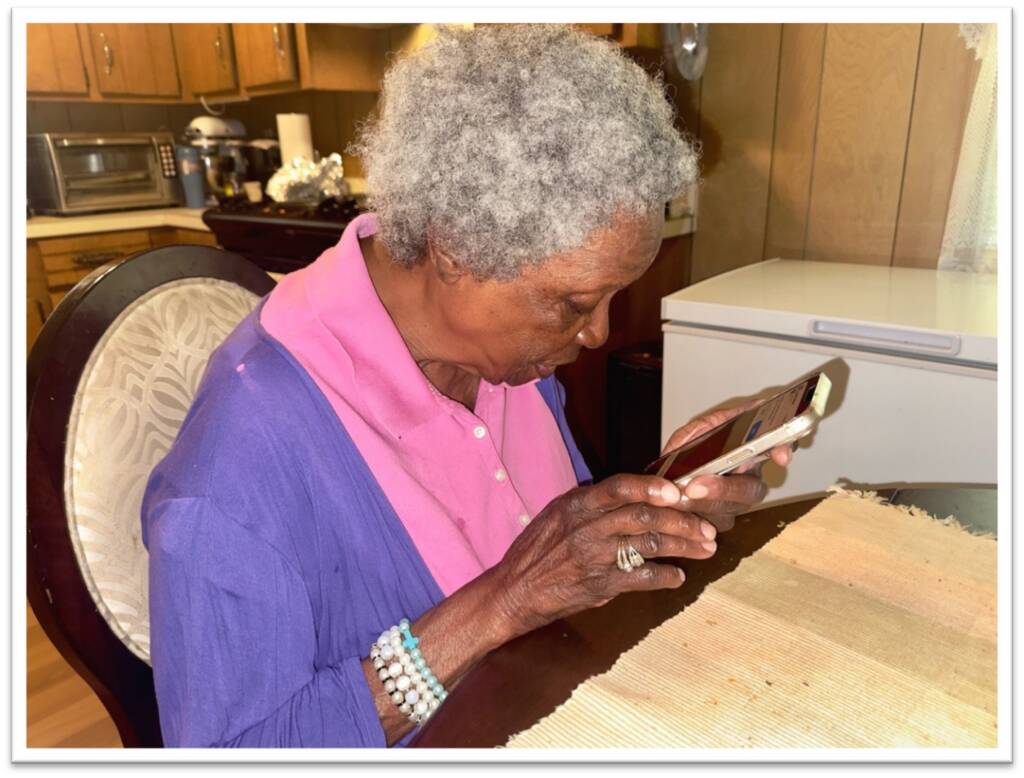 An elderly Black woman with curly grey hair looks down closely at a smartphone she is holding in her hands while sitting at a table.