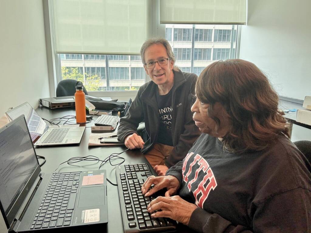iCanConnect trainer Harris Rosensweig smiles at the camera while participant Mitch Delaney types on a large-print external keyboard connected to a laptop during a training session.
