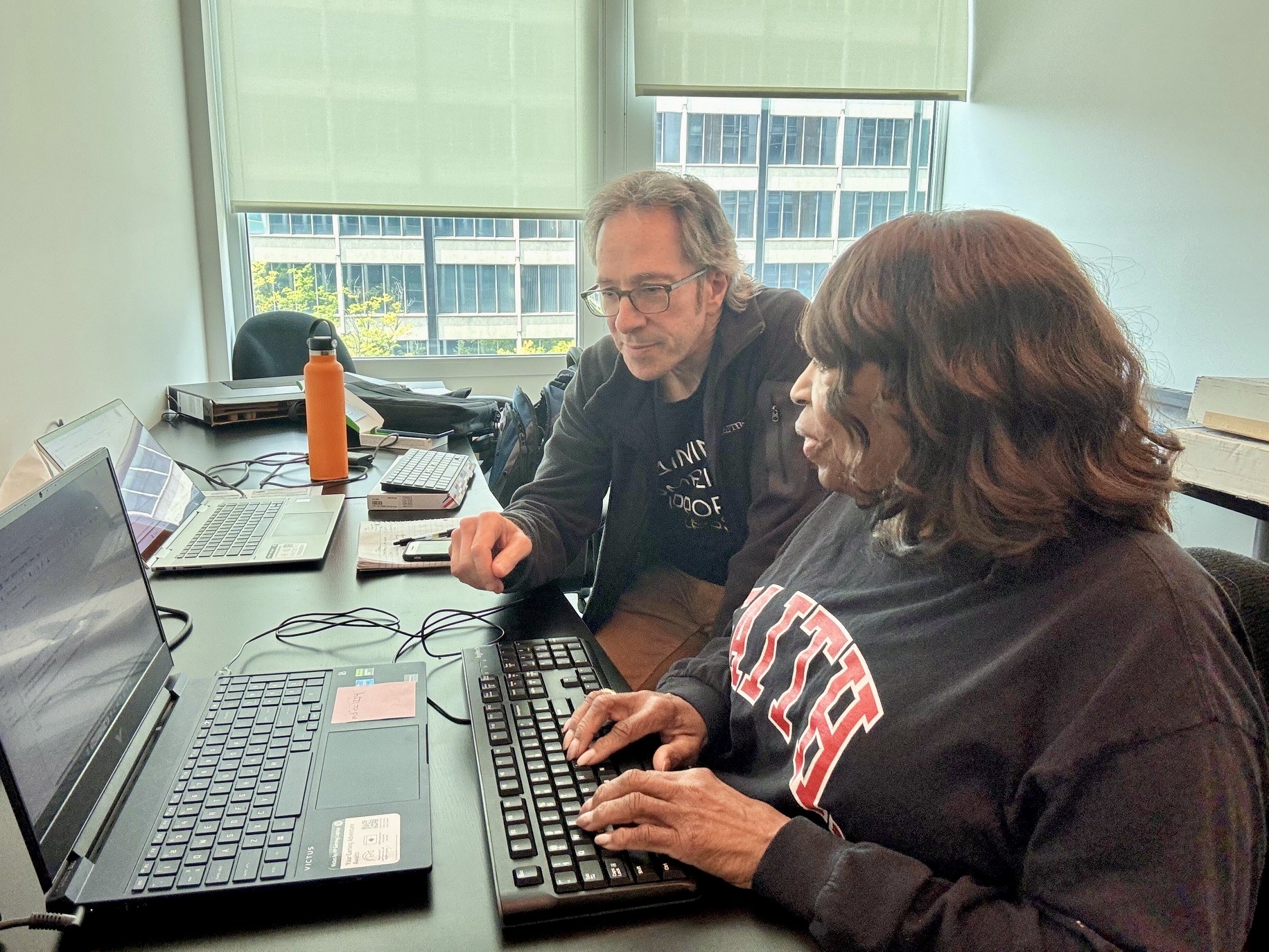 An iCanConnect trainer watches and points as a woman types on a large, high-contrast keyboard connected to a laptop in an office setting.